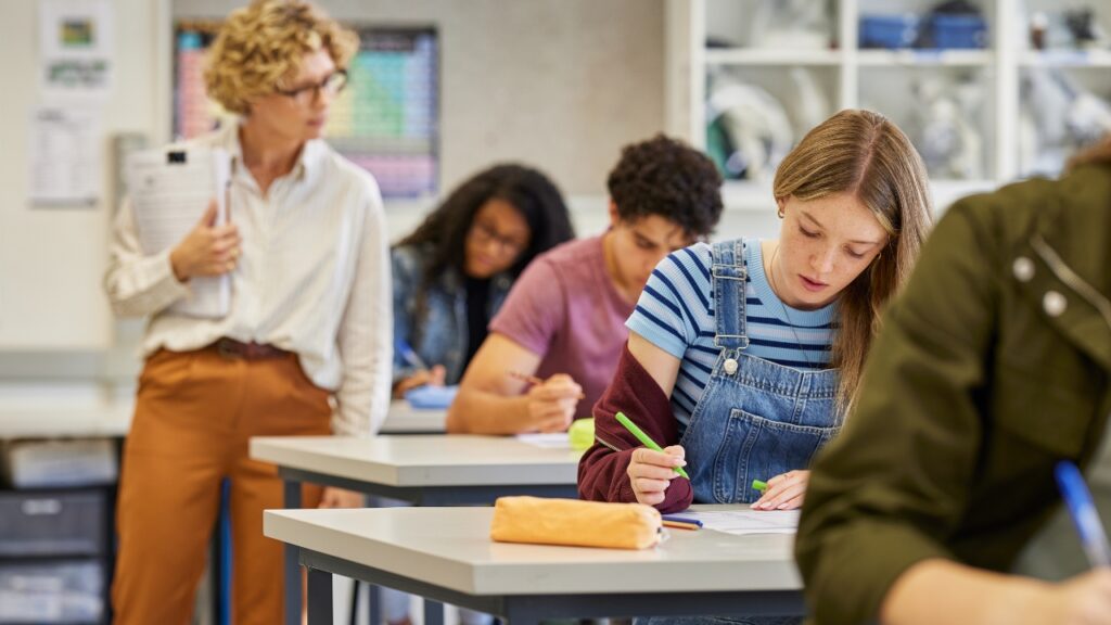 A focused student in overalls writes at a classroom desk with peers in the background. A teacher observes, holding papers, creating a studious atmosphere.