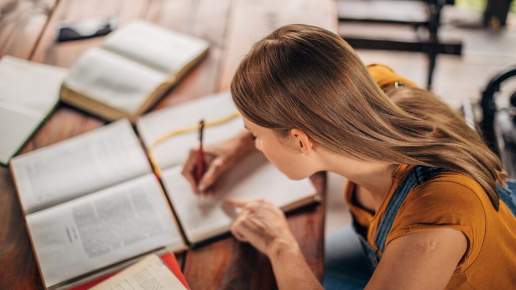 A young woman with brown hair studies at a wooden table, writing in a notebook. Open books surround her, suggesting a focused and studious environment.