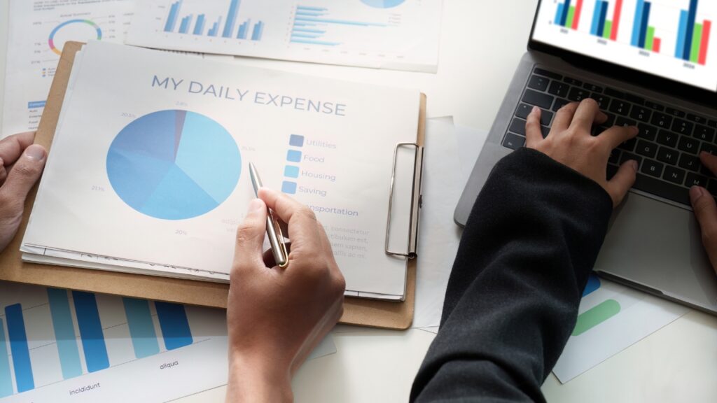 A person reviews a "My Daily Expense" pie chart on a clipboard, highlighting various categories. Another types on a laptop showing a bar graph. Professional tone.