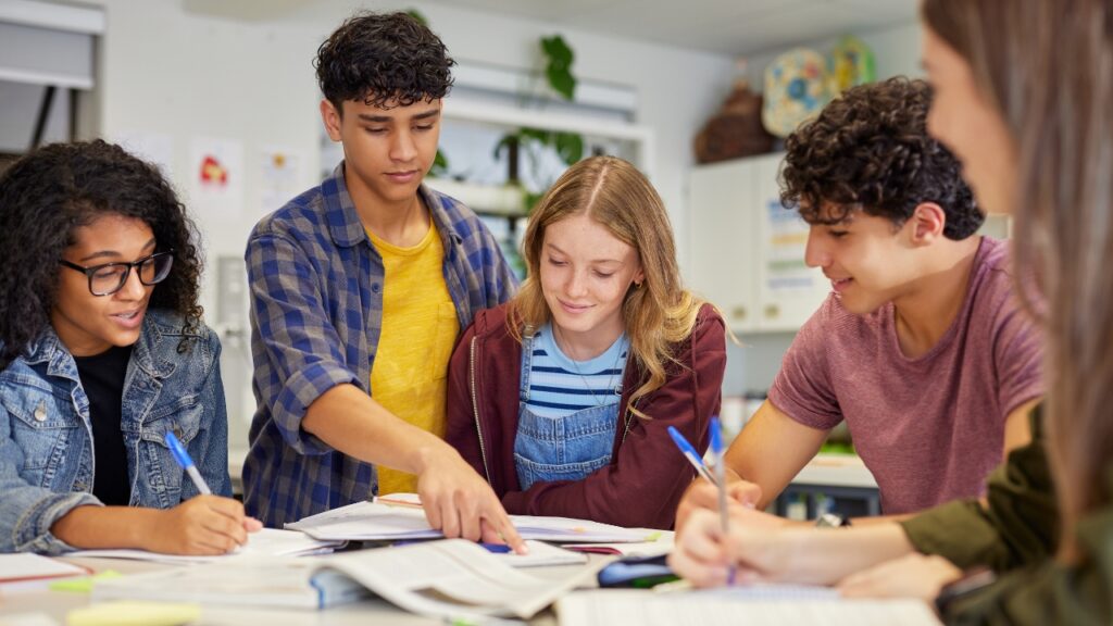A diverse group of five students collaborates around a table, engaged in discussion. One points at a book, suggesting teamwork in a lively classroom.
