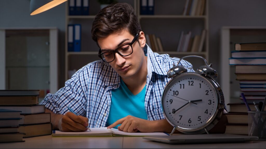 Young man with glasses studies late at a desk, surrounded by books, under a lamp's glow. A large clock shows 10:10 PM, highlighting a focused mood.
