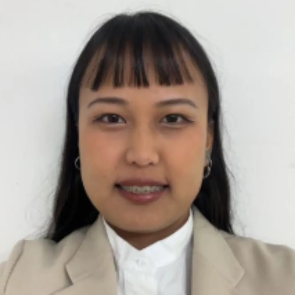 A person with long dark hair and fringe, smiling warmly. They are wearing a beige blazer and white shirt, set against a plain white background.