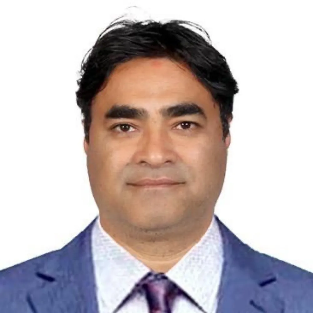 A man with short dark hair wearing a blue suit jacket, white shirt, and purple tie. He has a calm expression and a white background.