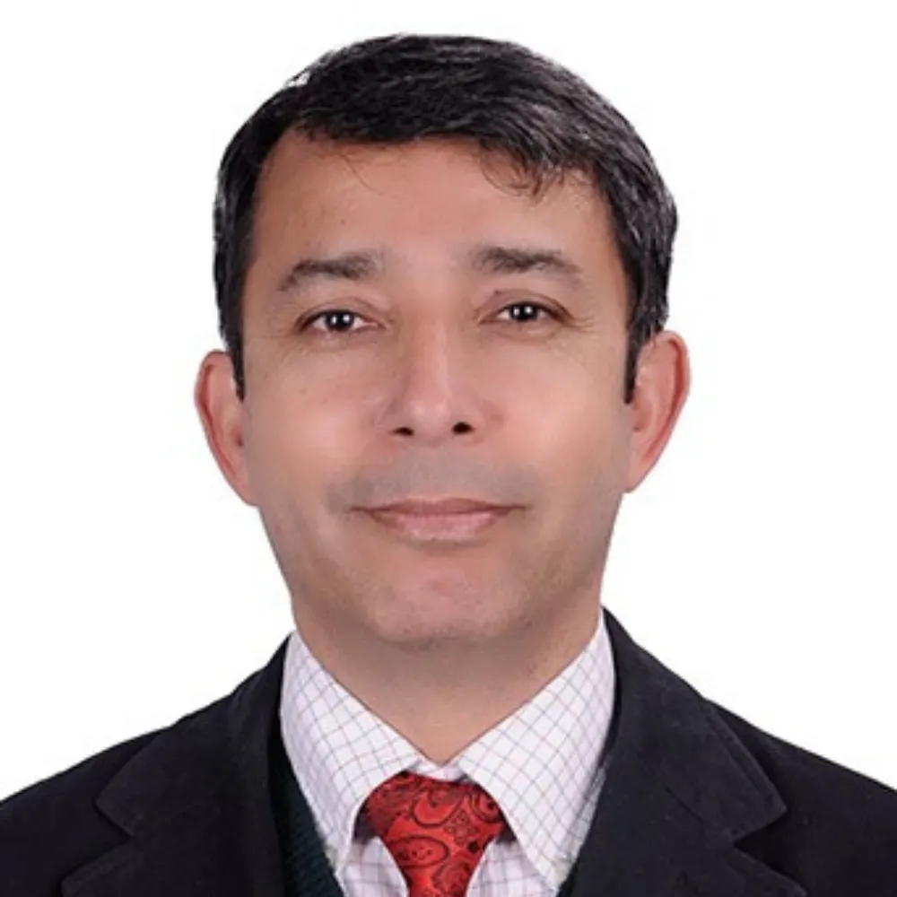 Portrait of a man in a formal suit with a checkered shirt and red tie. He has short, dark hair and a slight smile, set against a plain white background.