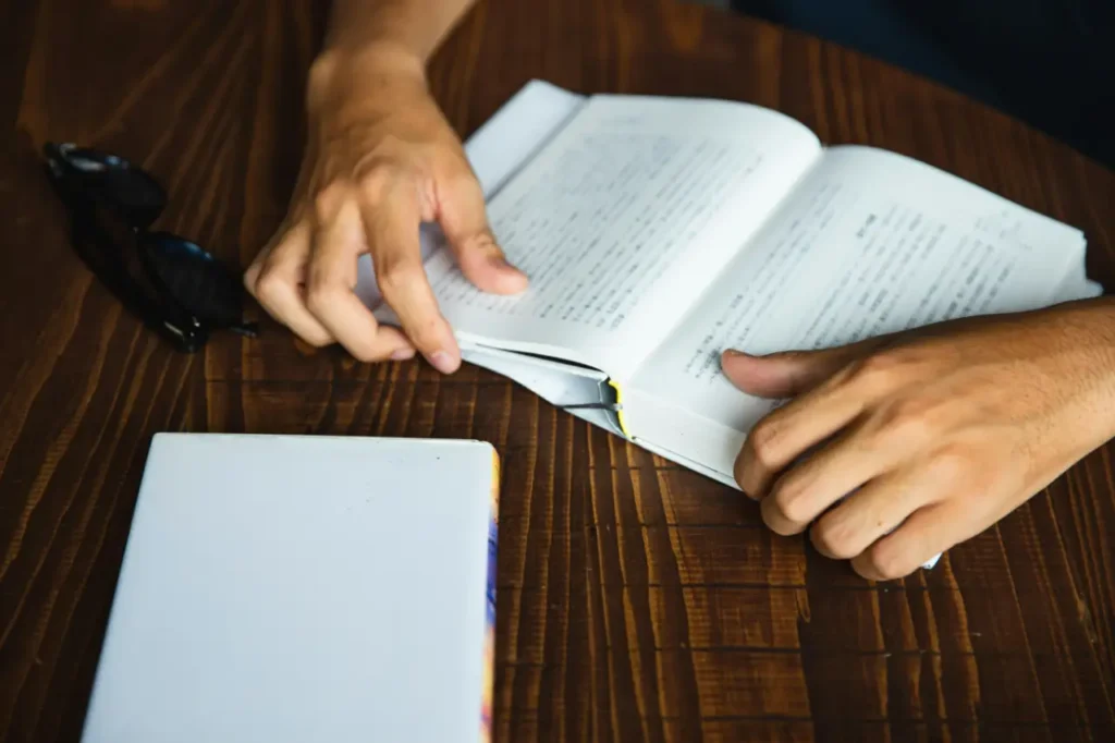 A close-up, high-angle shot of a person's tanned hands holding open a thick book with Japanese text on a dark, grained wooden table.
