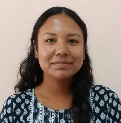 A head-and-shoulders portrait of a young woman with long, wavy dark hair and tan skin. She is looking directly at the camera with a subtle smile. She wears a navy blue top with a white, detailed block-print pattern and has small earrings. The background is a solid, light peach-colored wall.