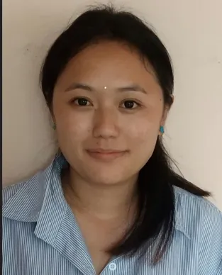 A portrait of a young woman with East Asian features, smiling slightly. She has dark hair tied back in a low ponytail and is wearing a light blue and white vertically striped button-down shirt. She has a small mark on her forehead and is wearing small turquoise earrings. The background is a plain, off-white wall.
