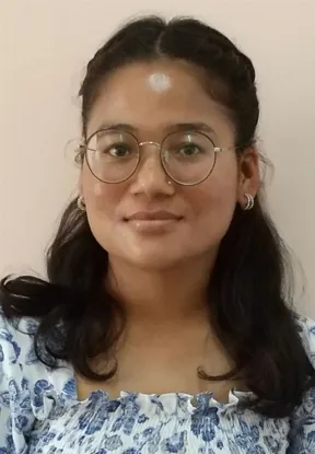 A close-up portrait of a young South Asian woman with long, dark hair parted in the middle and styled with small braids on top. She wears round, thin-rimmed glasses, small silver hoop earrings, and has a faint white circular mark on her forehead. She is wearing a blue and white floral smocked top against a plain, off-white background