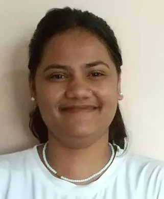 A headshot portrait of a smiling young woman with tan skin and dark hair pulled back. She is wearing a white t-shirt, a white beaded necklace, and small white stud earrings. The background is a plain, light-beige wall