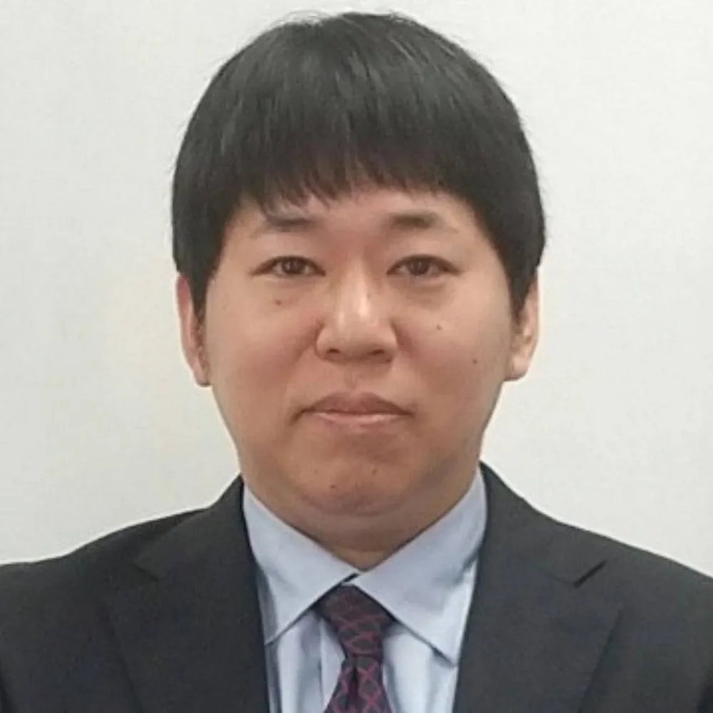 A man with short dark hair and a neutral expression is wearing a dark suit, light blue shirt, and patterned tie against a plain white background.