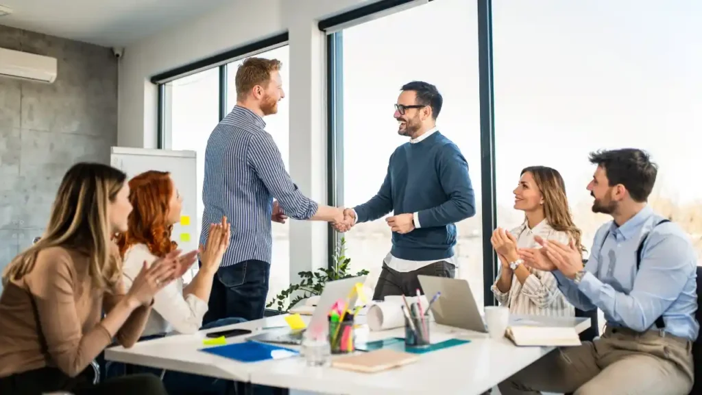 Two men shake hands in an office, while four colleagues sit around a table, clapping and smiling. The scene conveys success and teamwork.