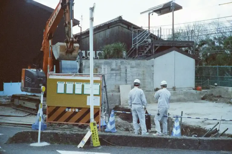 Two construction workers in hard hats stand near a caution sign at a building site. An excavator is digging beside them.